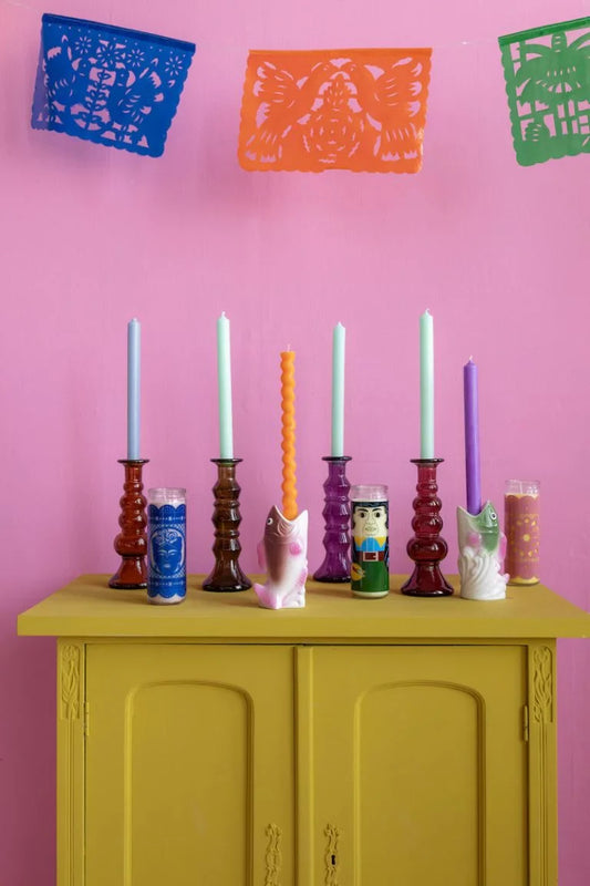 Colorful candles in various holders on a yellow cabinet against a pink wall with colorful cut-out decorations.