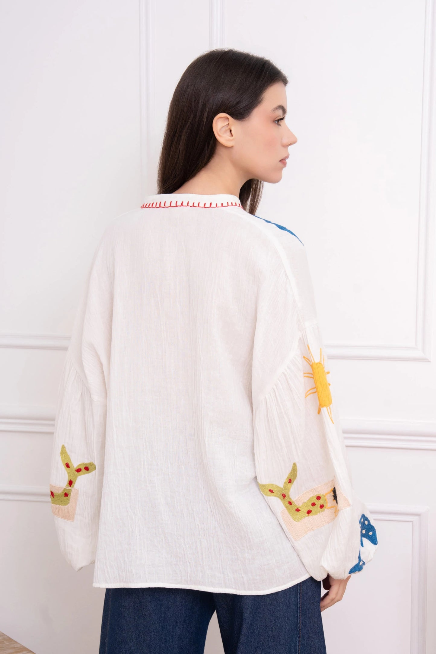 Woman wearing a white cardigan with colorful patterns, standing against a white wall.