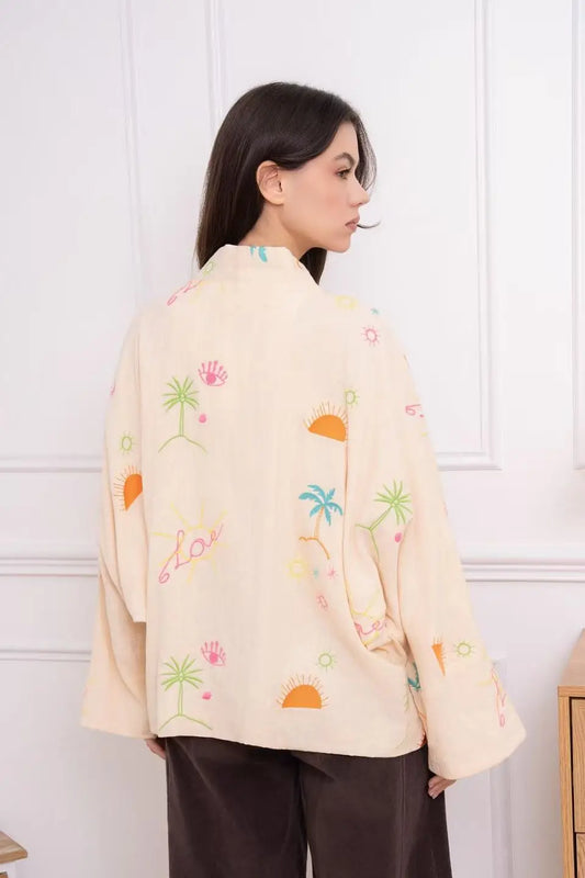 Woman wearing a light-colored jacket with colorful patterns against a white wall.
