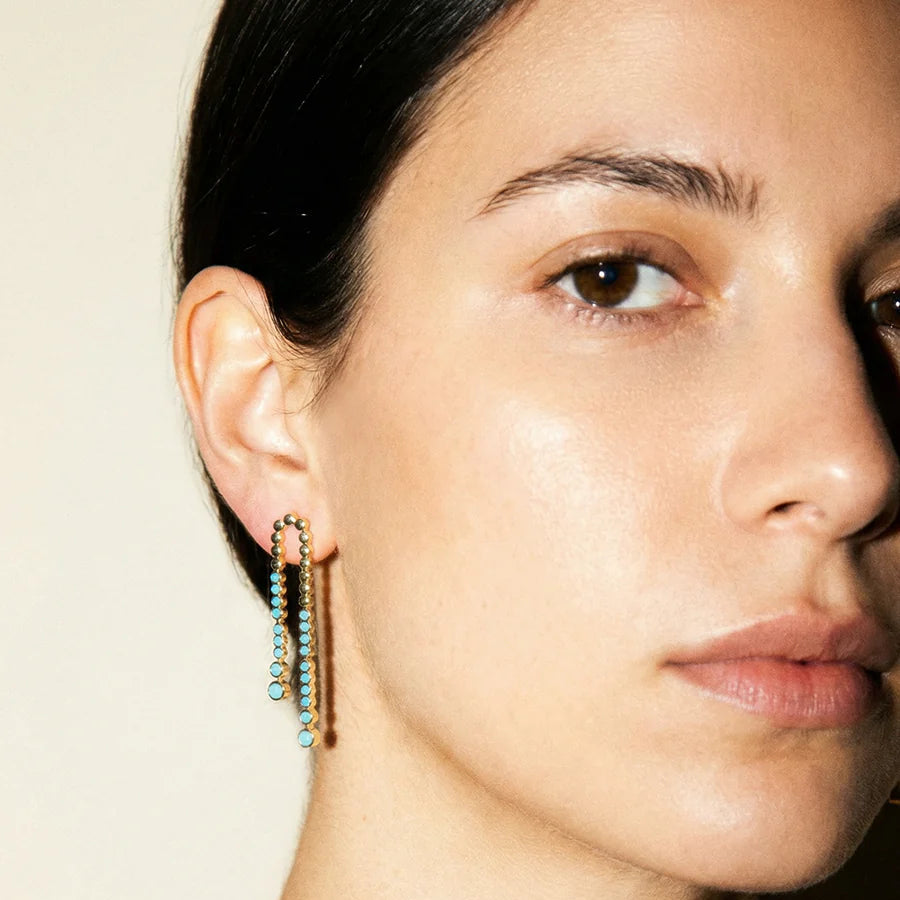 Close-up of a woman wearing gold earrings with green beads on a neutral background