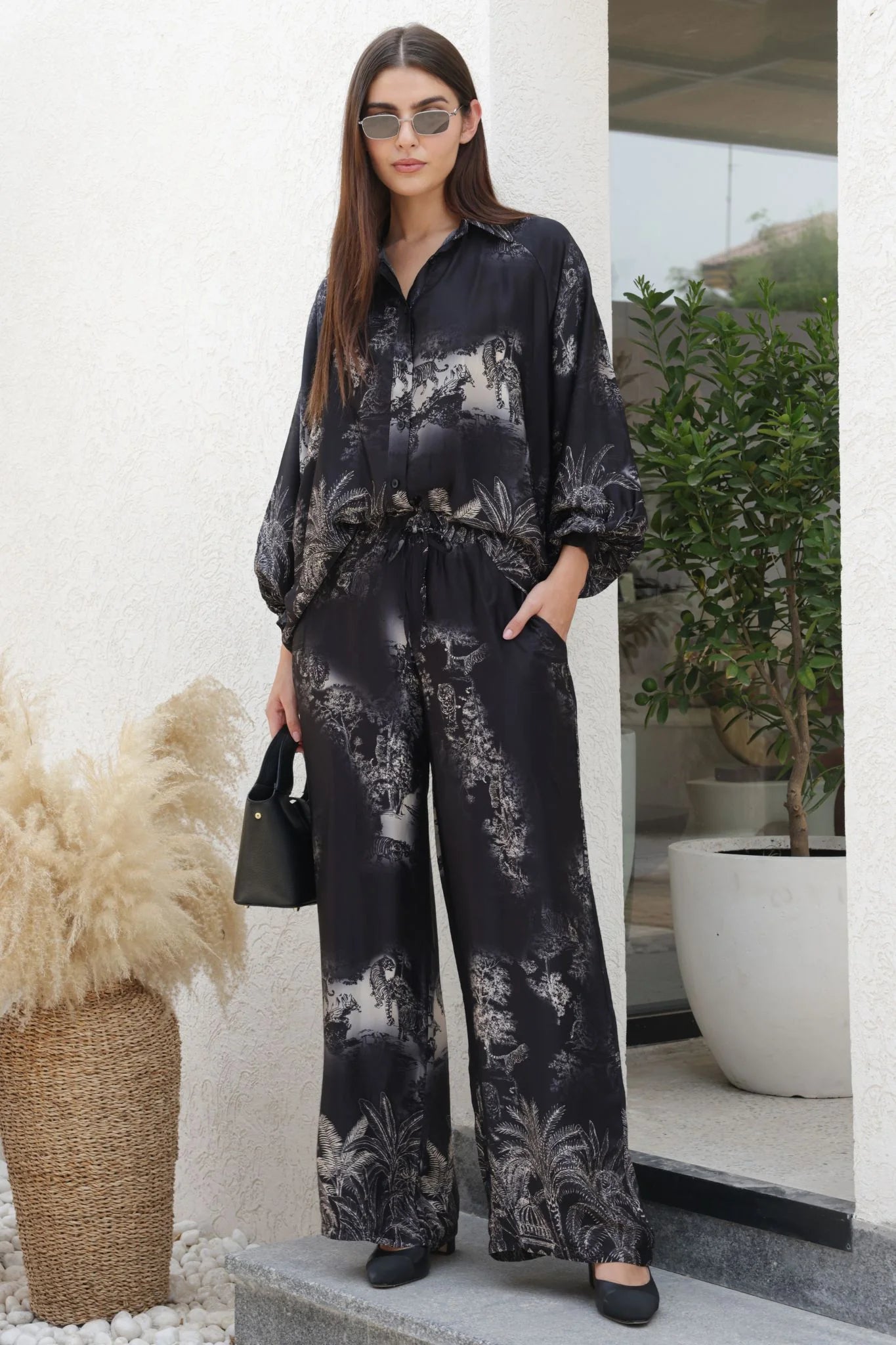 Woman wearing a black floral outfit standing outdoors with plants and a building in the background.