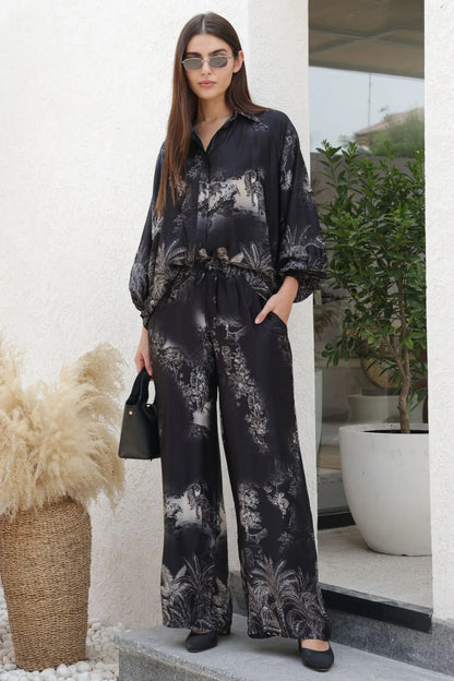 Woman wearing a black floral outfit standing outdoors with plants and a building in the background.