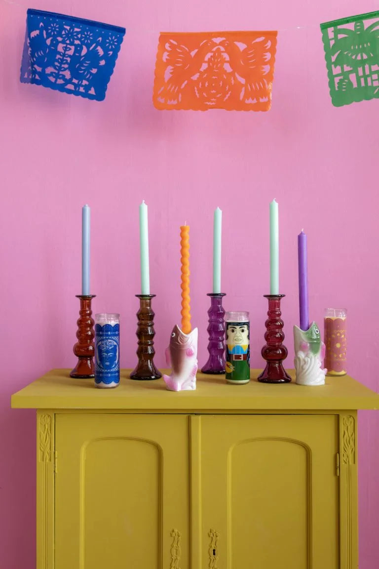 Colorful candles in various holders on a yellow cabinet against a pink wall with colorful cut-out decorations.