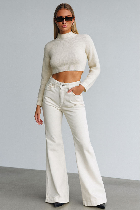 Woman wearing a white cropped sweater and white flared jeans against a gray background