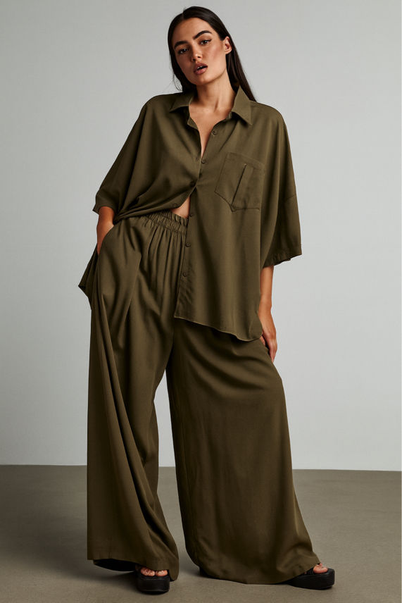 Woman wearing an olive green outfit with wide-leg pants and a shirt against a plain background