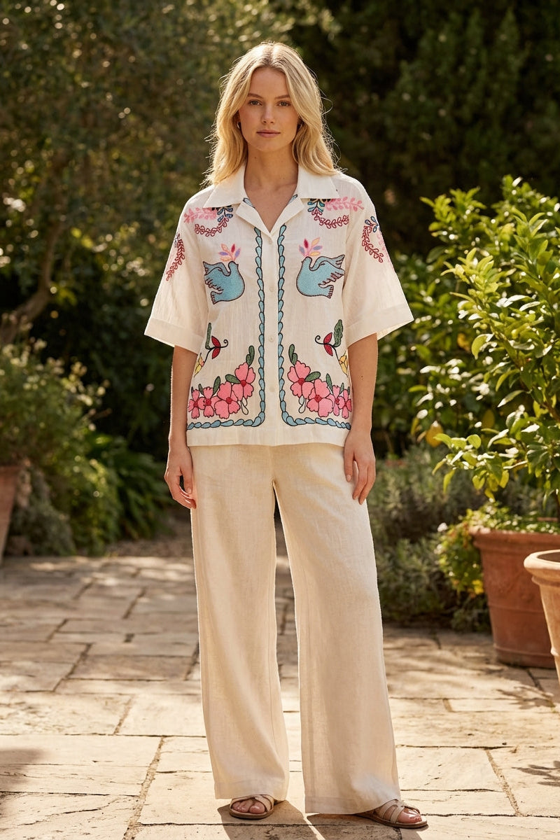 Woman wearing a patterned shirt and beige pants standing outdoors with greenery in the background