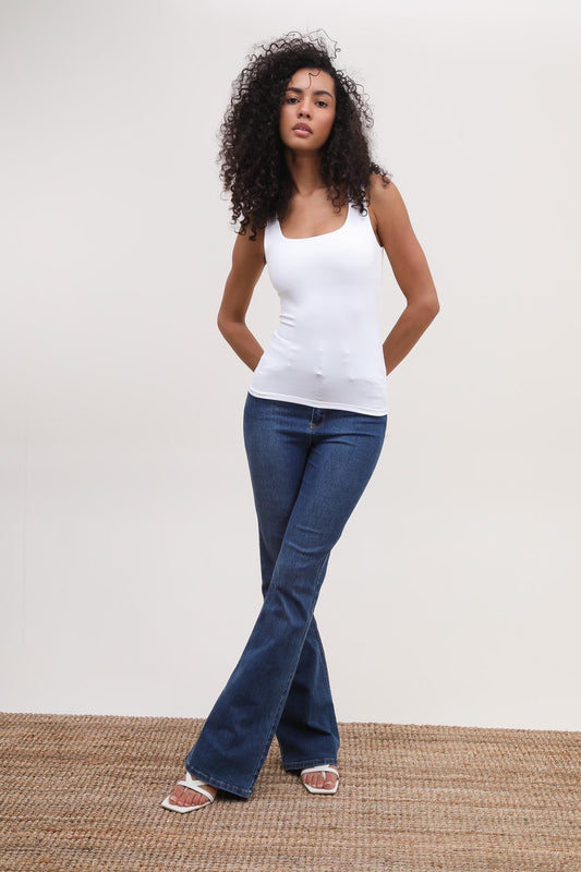 Woman wearing a white tank top and blue jeans standing on a woven mat with a plain background