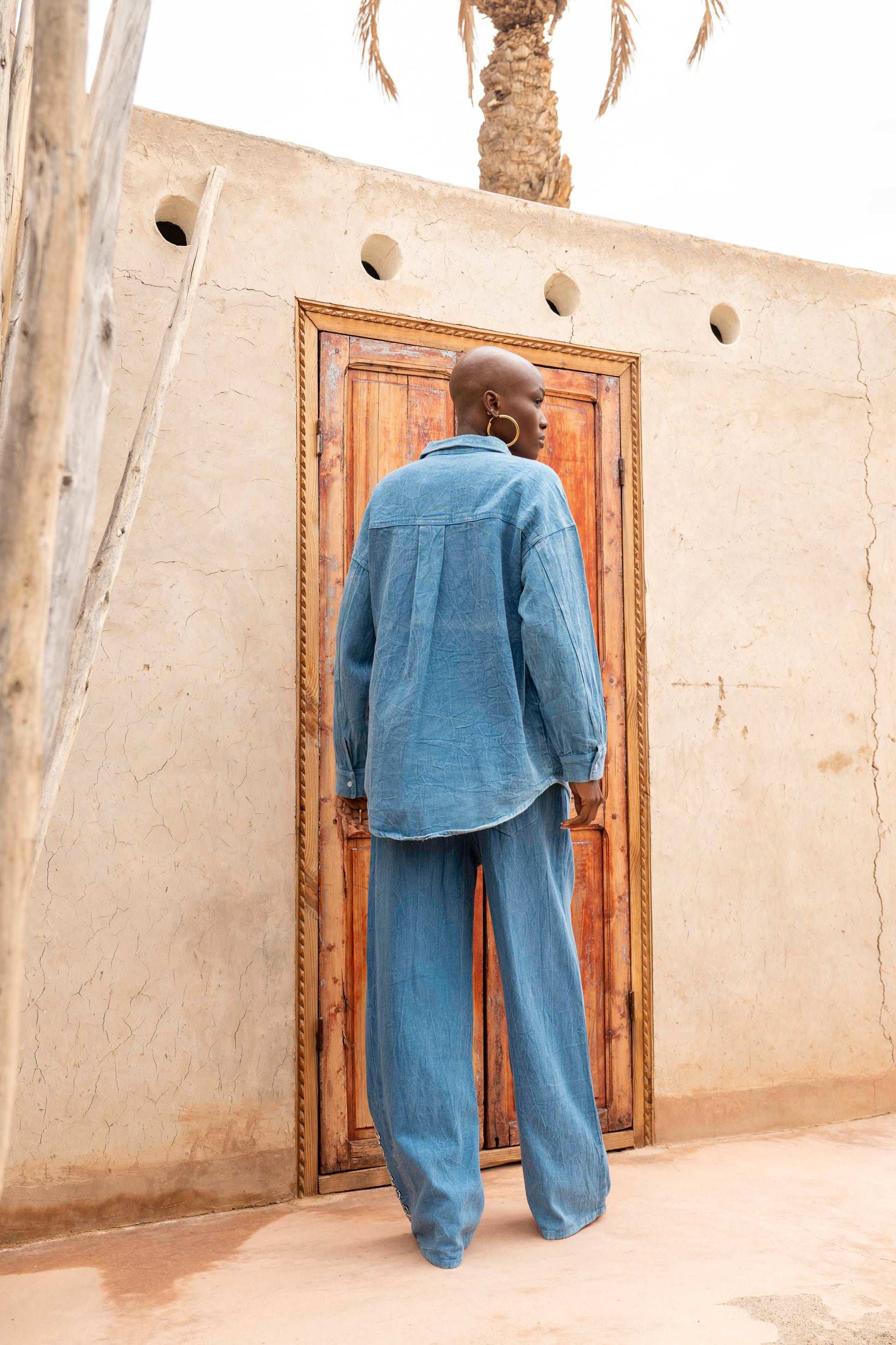 Man in blue outfit standing in front of a wooden door on a beige wall with a palm tree in the background.
