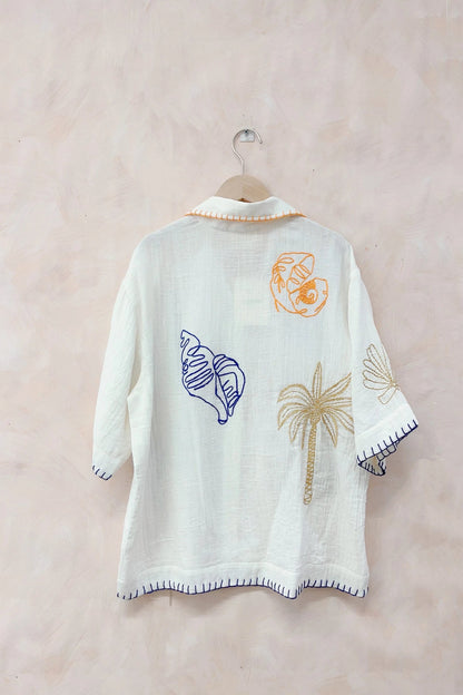 White embroidered poncho with shell and palm tree designs on a beige background