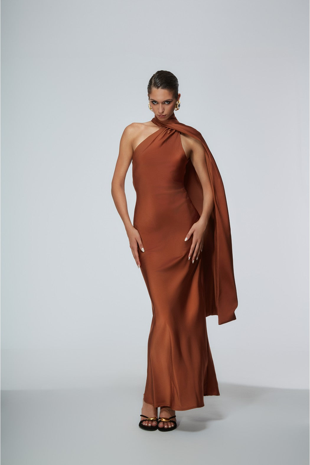 Woman wearing a rust-colored one-shoulder gown with a cape on a white background