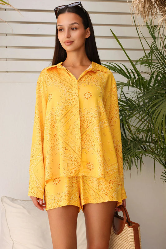 Woman wearing a yellow outfit with a pattern, standing outdoors with plants in the background.