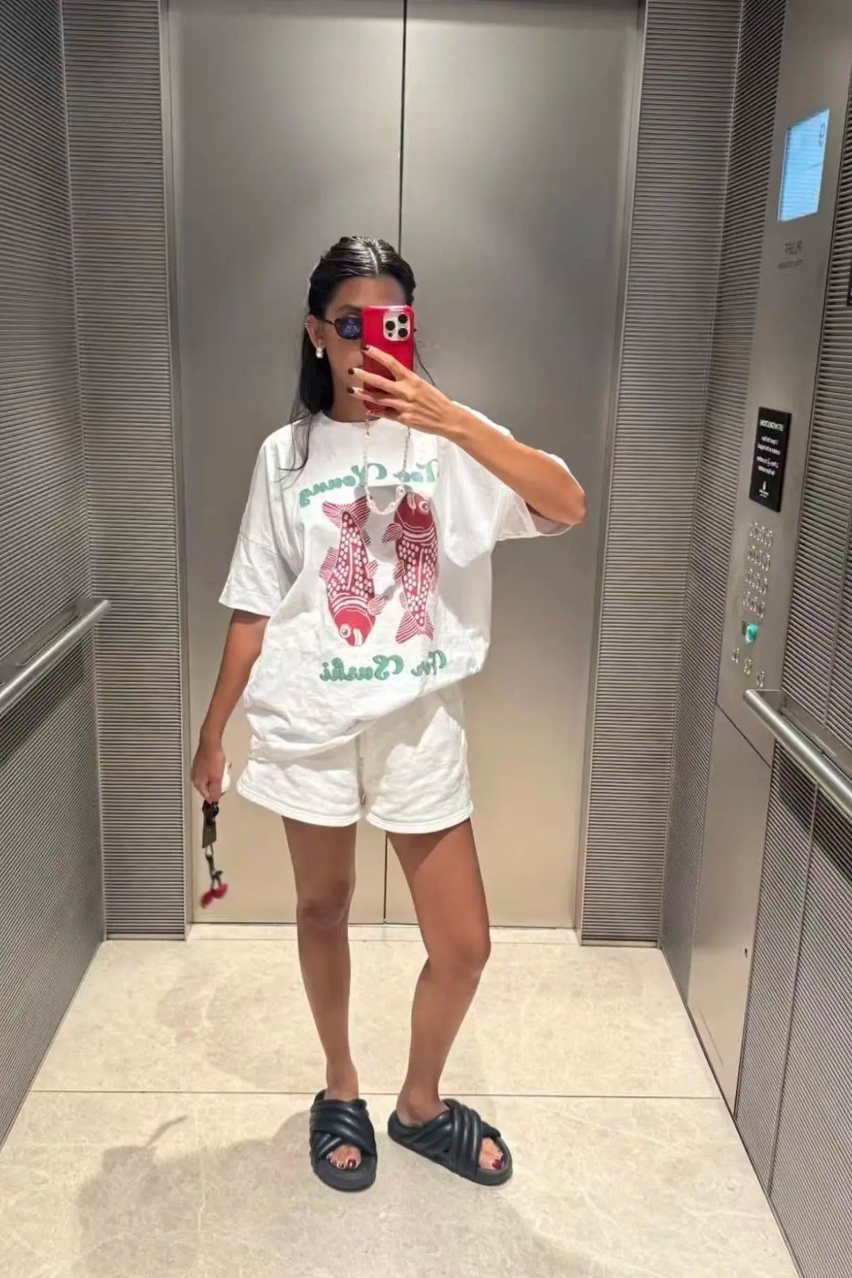 Person taking a mirror selfie wearing a white t-shirt with graphics and white shorts in an elevator.