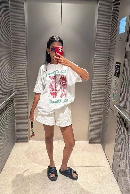 Person taking a mirror selfie wearing a white t-shirt with graphics and white shorts in an elevator.