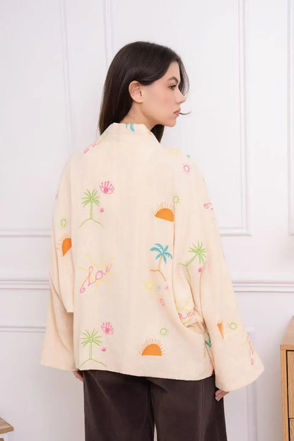 Woman wearing a light-colored jacket with colorful patterns against a white wall.
